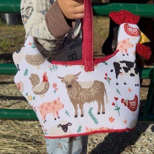 May include: A fabric basket shaped like a chicken with a red handle and trim. The white exterior features a repeating pattern of farm animals including a brown highland cow, a pink pig, a black and white cow, sheep, and chickens with small flowers.