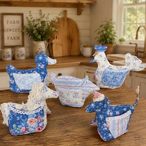 May include: A collection of four fabric animal figures including a blue floral duck, a white floral rooster, a horse with a floral mane, and a blue floral cow. Each stuffed animal features a small pocket on its side with white lace trim.