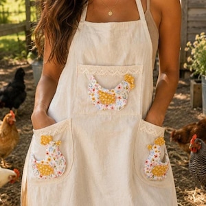 May include: A cream-coloured apron with two pockets and a central pocket, all decorated with a floral chicken applique. The apron has lace trim on the pockets and top edge. The chicken appliques have yellow and floral patterns.