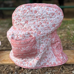 May include: A quilted fabric cover for a kitchen appliance featuring a pink and white floral and fruit pattern. The cover has a rounded top, a structured shape, and a small rectangular pocket on the front with a horizontal striped design.