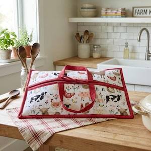 May include: A rectangular fabric casserole carrier with a white background featuring a colourful farm animal print including cows, pigs, sheep, and chickens. The carrier has red fabric trim, red handles, and a central tie closure on the top.