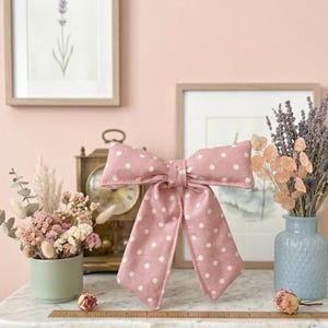 May include: A large, pink fabric bow with white polka dots. The bow is the focal point, set against a backdrop of framed art, a vintage clock, and floral arrangements. The bow's dimensions are approximately 12 inches wide.