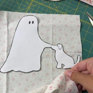 May include: A white fabric ghost cutout with black eyes and a black outline, positioned next to a white fabric dog cutout with a black outline, both resting on a light-coloured fabric background featuring a small pink floral pattern.
