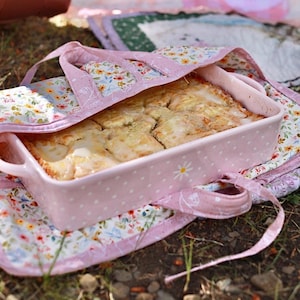 May include: A rectangular pink ceramic baking dish with white polka dots and side handles, containing a baked pastry with a light glaze. The dish is nestled inside a fabric carrier featuring a floral pattern on a pink and white background.