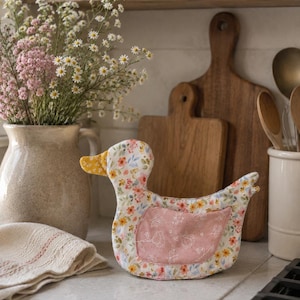 Pode incluir: Um acessório de cozinha em tecido com formato de pato, apresentando uma base branca com um padrão floral colorido de pequenas flores rosas, amarelas e azuis. O pato tem um bico amarelo sólido e um painel central de tecido rosa seco.