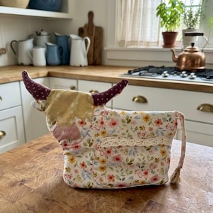 Peut inclure: Une théière en tissu en forme de vache avec un motif floral dans des tons rose, jaune et vert. La vache a des cornes bordeaux foncé avec un petit motif blanc, une tache de tête couleur moutarde et une bordure en dentelle crème sur le côté.