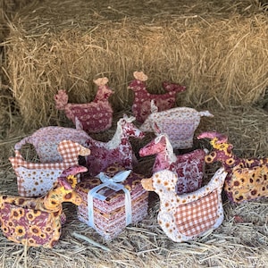 May include: A collection of fabric animal figures including chickens, horses, and ducks, featuring various patterns such as sunflowers, orange and white gingham, and floral prints, arranged around a square stack of fabric squares tied with a white ribbon.