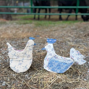 May include: Two fabric chicken-shaped ornaments. The left one features a beige and blue striped pattern with floral accents. The right one displays a white fabric with blue floral prints, a blue patterned comb, and a lace-trimmed pocket.