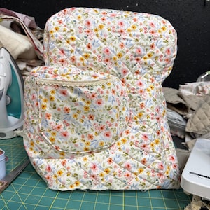 Puede incluir: Una funda de tela acolchada para electrodomésticos de cocina con fondo blanco y un patrón floral repetitivo de pequeñas flores rosas, naranjas y amarillas con hojas verdes. La funda incluye un bolsillo frontal redondeado con borde de encaje crema.