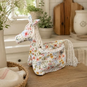 May include: A stuffed horse toy made of white fabric with a colourful floral pattern featuring pink, yellow, and green flowers. The horse has a cream-coloured yarn mane and tail, and a small pocket on its side trimmed with white lace.