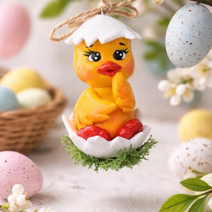 Puede incluir: Adorno de pollito amarillo con sombrero de cáscara de huevo blanco y pies rojos, suspendido de una cuerda. El pollito está en una base de cáscara de huevo blanca con musgo verde. Huevos de Pascua y flores en tonos pastel en el fondo.
