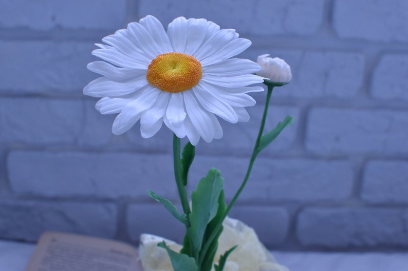 May include: A single white daisy with a yellow centre and a bud on a green stem. The flower is in focus against a blurry background of a white brick wall.