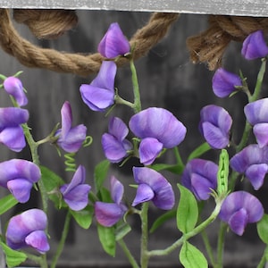 May include: A cluster of purple sweet pea flowers with green leaves and stems arranged in a rustic wooden frame.