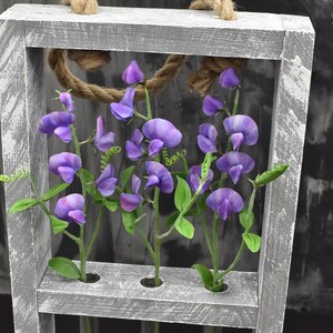 May include: A gray wooden frame with four small glass tubes, each holding a stem of purple sweet pea flowers. The frame is hanging from a rope and has a distressed white paint finish.