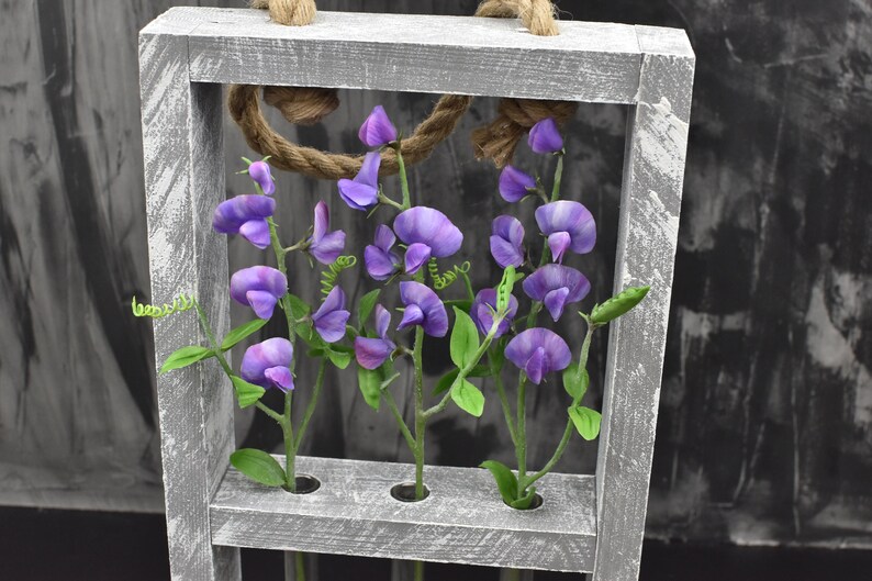 May include: A gray wooden frame with four small glass tubes, each holding a stem of purple sweet pea flowers. The frame is hanging from a rope and has a distressed white paint finish.