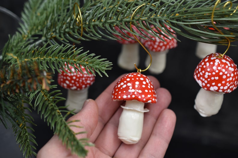 May include: A set of four red and white mushroom-shaped Christmas ornaments with gold wire hangers. The ornaments are made of polymer clay and have a white stem and a red cap with white dots.