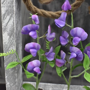 May include: Purple sweet pea flowers with green stems and leaves against a gray wooden background.