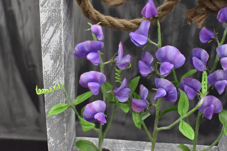 May include: Purple sweet pea flowers with green stems and leaves against a gray wooden background.