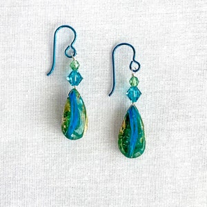Op de afbeelding: Een paar druppelvormige oorbellen met blauwe en groene gemarmerde stenen accenten. De oorbellen zijn gemaakt met titanium haakjes en hebben kristallen kralen in tinten groen en blauw.