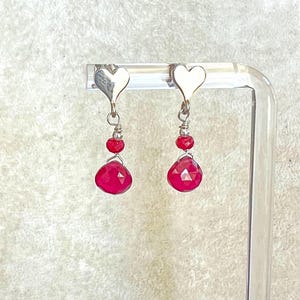 May include: A pair of silver earrings with heart-shaped tops. Each earring features a small red bead, a cluster of two smaller red beads, and a large, faceted, teardrop-shaped red gemstone. The earrings are displayed on a clear stand.