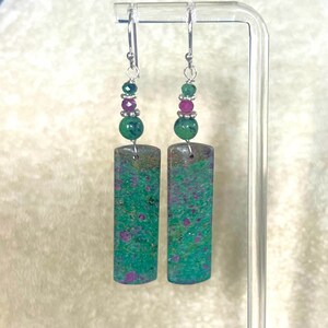 May include: A pair of dangle earrings with a rectangular, green and purple stone pendant. The earrings have a silver hook and are adorned with green and purple beads. The earrings are displayed on a clear stand.