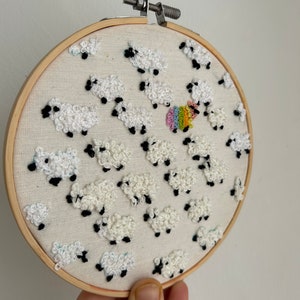 May include: Embroidered hoop art featuring a white sheep with a rainbow design, surrounded by other white sheep. The sheep are embroidered with white yarn and have black eyes.