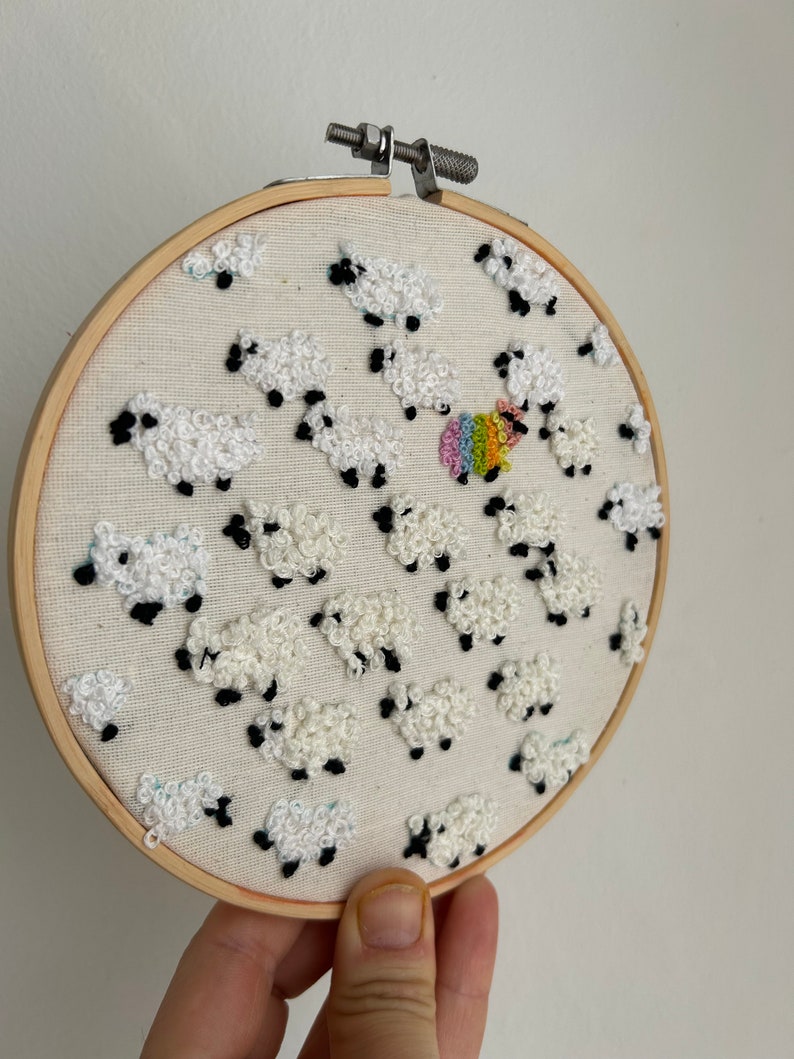 May include: Embroidered hoop art featuring a white sheep with a rainbow design, surrounded by other white sheep. The sheep are embroidered with white yarn and have black eyes.