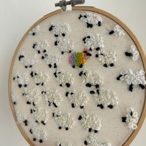 May include: A white embroidery hoop with a design of white sheep with black eyes and legs. The sheep are arranged in a circular pattern around a rainbow colored circle in the center.