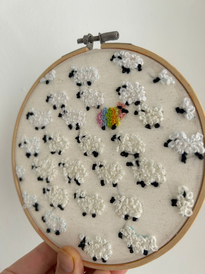 May include: A white embroidery hoop with a design of white sheep with black eyes and legs. The sheep are arranged in a circular pattern around a rainbow colored circle in the center.