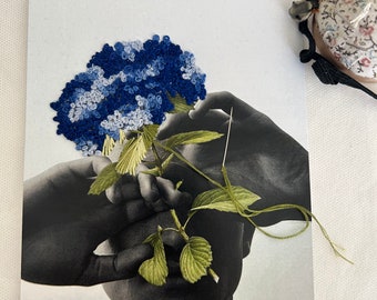 May include: A black and white photograph of a person's face with a blue and white hydrangea flower covering their eyes. The flower is made of fabric and is attached to a green stem with leaves.