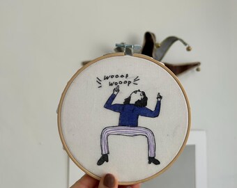 May include: Embroidered hoop art featuring a cartoon person with arms raised in the air, shouting "wooop wooop" in black text. The person is wearing a blue and white striped shirt and purple pants.