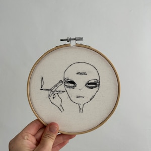 Könnte beinhalten: Stickerei auf einem runden Rahmen mit einer Schwarzweißillustration eines Außerirdischen, der eine Zigarette raucht.