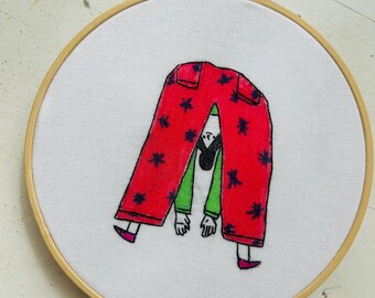 May include: Embroidered hoop art featuring a person with a green shirt and red pants with black stars. The person is upside down with their legs in the air.
