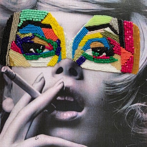 May include: A black and white photograph of a woman wearing a colourful beaded mask over her eyes. She is holding a cigarette in her hand and her mouth is slightly open.