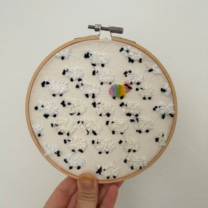 May include: A white embroidery hoop with a design of white sheep with black faces. One sheep is rainbow colored.