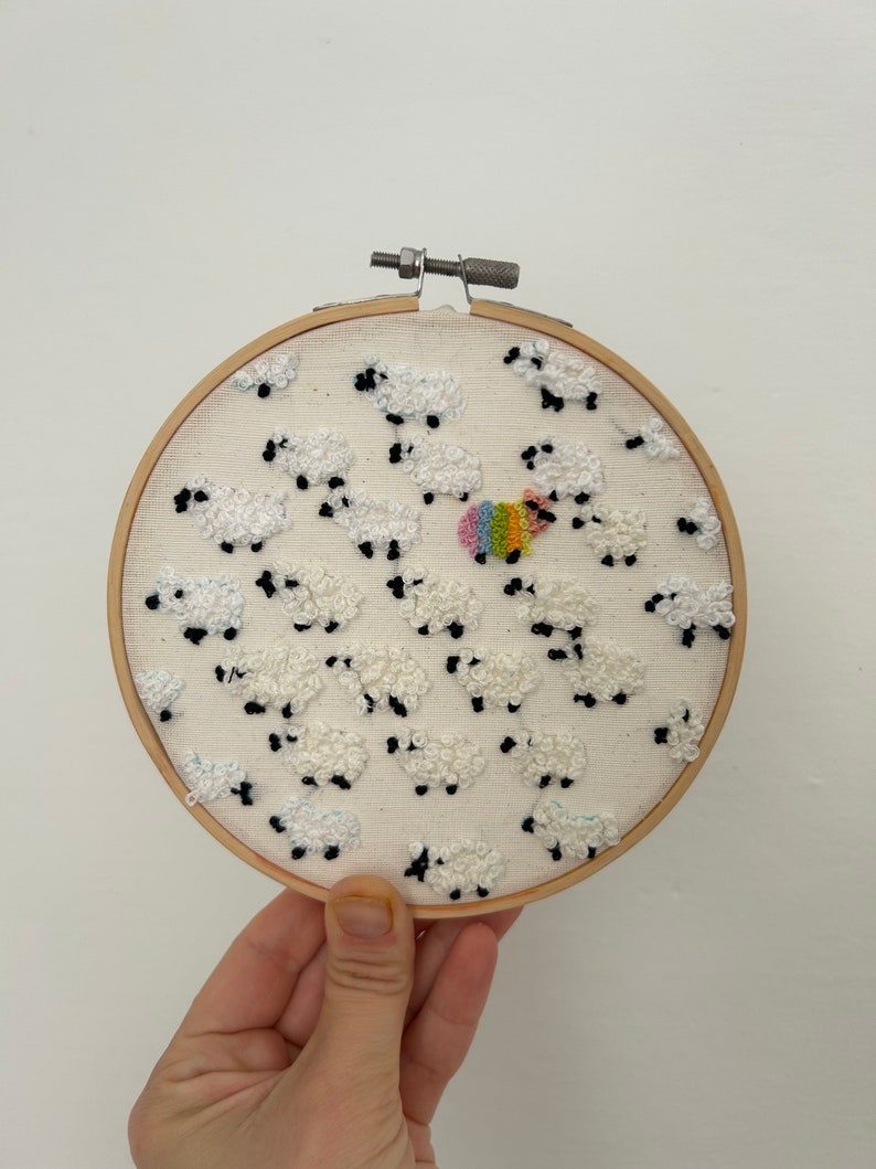 May include: A white embroidery hoop with a design of white sheep with black faces. One sheep is rainbow colored.