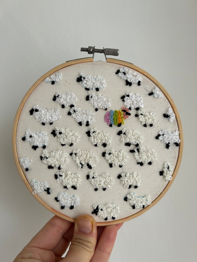 May include: Embroidered hoop art featuring a white sheep with a rainbow fleece surrounded by white sheep with black eyes and hooves.