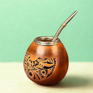 Puede incluir: Una taza de mate de calabaza marrón con un diseño decorativo negro y naranja. Un borde de metal plateado rodea la abertura. Una pajita de metal plateado con un anillo dorado está insertada en la taza. El fondo es de color verde claro.
