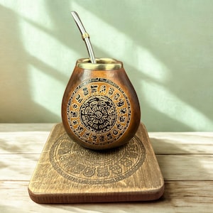 Puede incluir: Una taza de mate de calabaza marrón con una pajita de metal, con un diseño circular negro y marrón detallado. La taza se encuentra sobre un posavasos de madera con un patrón grabado a juego. Un recipiente tradicional sudamericano.