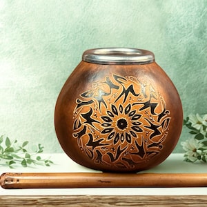 Puede incluir: Una taza de mate marrón en forma de calabaza con un borde plateado y un diseño decorativo negro y marrón. Una pajita de madera, o bombilla, descansa frente a la taza. La taza es un recipiente tradicional para beber yerba mate.