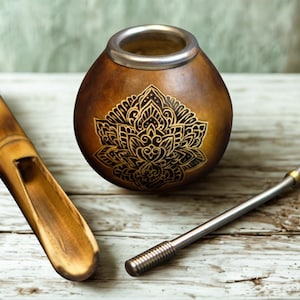 Puede incluir: Una calabaza marrón con borde plateado y diseño floral dorado, una cuchara de madera y una pajita de metal descansan sobre una superficie de madera. La calabaza se utiliza para beber yerba mate, una bebida tradicional sudamericana.