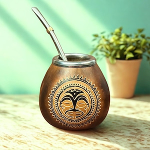 Puede incluir: Una taza de mate de calabaza marrón con borde plateado y pajita de metal. La taza presenta un diseño de rostro tribal tallado en negro y dorado. Una pequeña planta en una maceta beige está en el fondo.