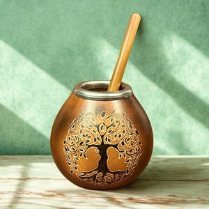 Puede incluir: Una taza de mate de calabaza marrón con borde plateado y pajita de madera. La taza presenta un diseño detallado de árbol de la vida. La pajita está insertada en la taza. El fondo es de color verde claro.