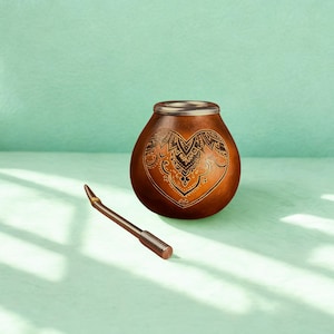 Puede incluir: Una taza de yerba mate en forma de calabaza marrón con un diseño de corazón decorativo y un borde de metal. Una pajita de metal, o bombilla, descansa junto a la taza. El fondo es de color verde claro.
