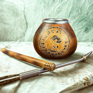 Puede incluir: Una taza de mate de calabaza marrón con un borde plateado y un diseño tallado detallado. Una pajita de madera y una pajita de metal se colocan frente a la taza. El fondo es una tela estampada en verde claro y blanco.