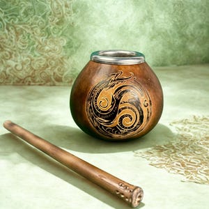 Puede incluir: Una taza de mate de calabaza marrón con borde plateado y diseño de dragón tallado. Una pajita de madera, o bombilla, descansa junto a la taza. El fondo es verde claro con un patrón decorativo.