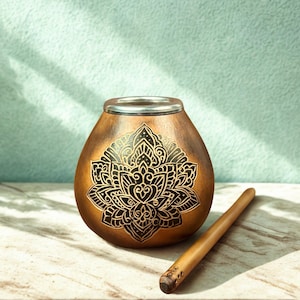Puede incluir: Una taza de yerba mate en forma de calabaza marrón con un borde plateado y un diseño floral negro detallado. Una pajita de madera, o bombilla, descansa junto a la taza. El fondo es de color verde azulado claro.