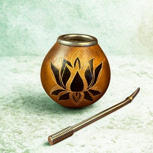 Puede incluir: Una taza de mate de calabaza marrón con borde plateado y diseño de flor de loto negra. Una pajita bombilla de metal descansa junto a la taza. La calabaza mide unos 10 cm de altura.