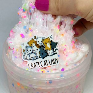 May include: White slime with colorful sprinkles in a clear plastic container. The slime is being pulled out of the container with a finger. A sticker with a group of cartoon cats and the text "Crazy Cat Lady" is on the top of the slime.