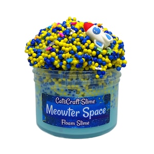 May include: A clear plastic container filled with blue, yellow, and white foam slime. The slime is topped with yellow and blue beads, star-shaped sprinkles, and a small white rocket ship. The container has the text "CatsCraft Slime Meowter Space Foam Slime".